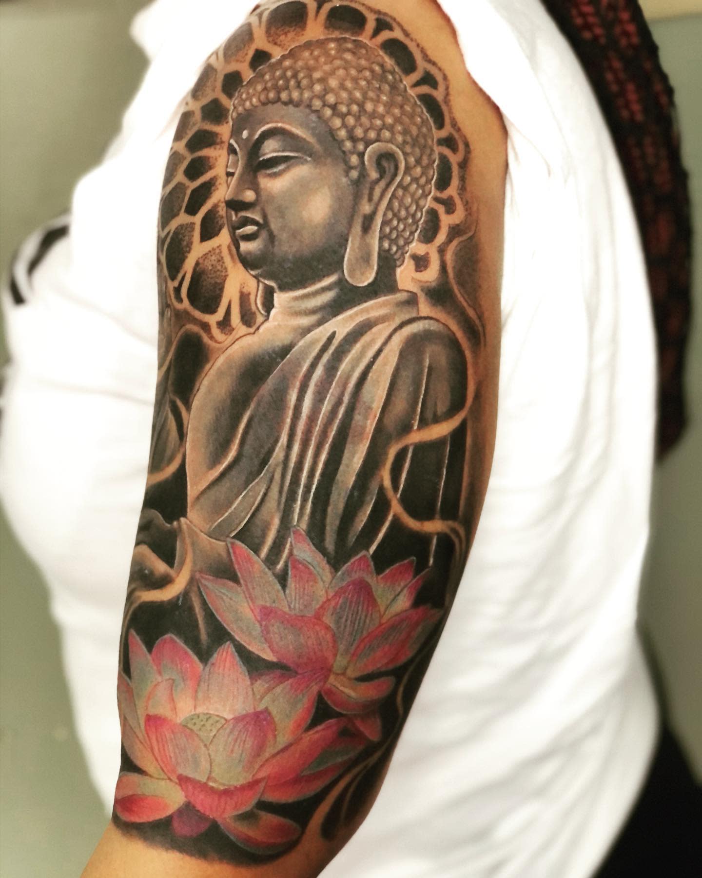 Buddha tattoo on upper arm with lotus flowers in black, grey, and pink